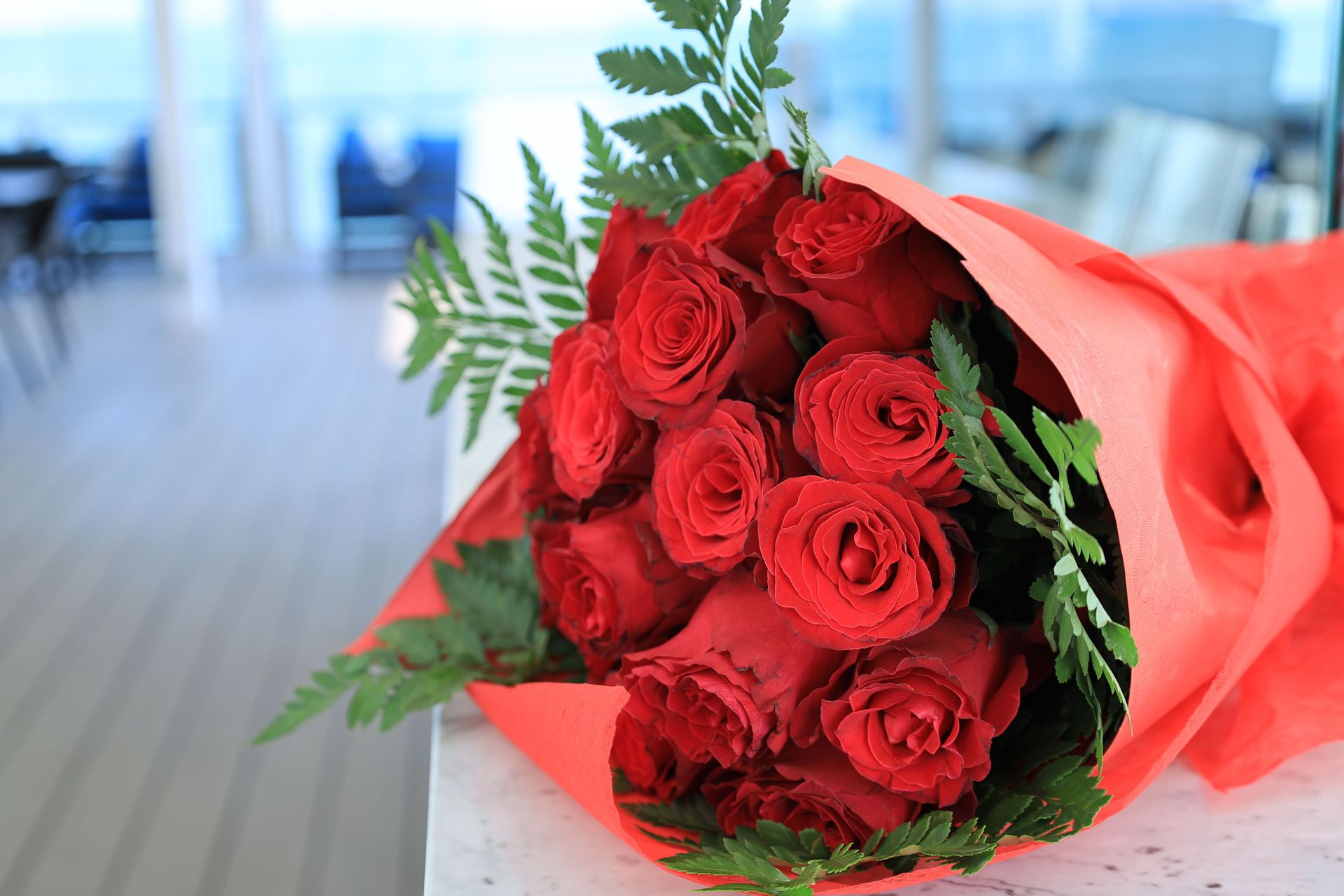 Close-up of a vivid red rose bouquet wrapped in orange paper with green leaves indoors.